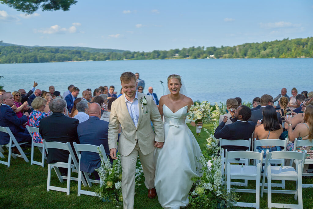 Newlyweds walk up the aisle after a lakeside ceremony as guests applaud.
