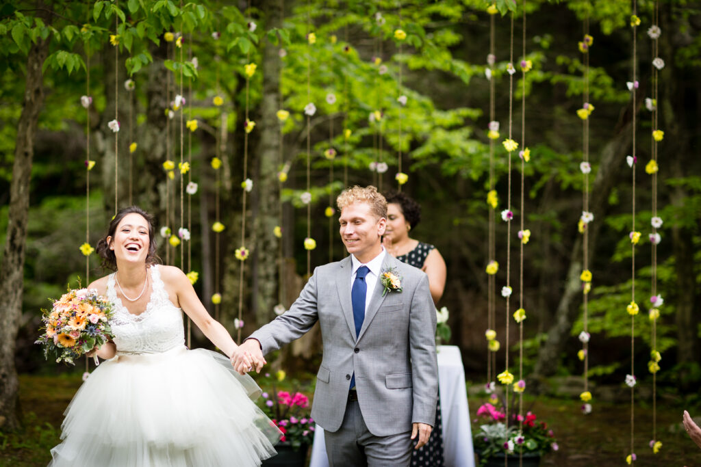 Laughing newlyweds hold hands beneath hanging flowers after their outdoor ceremony.