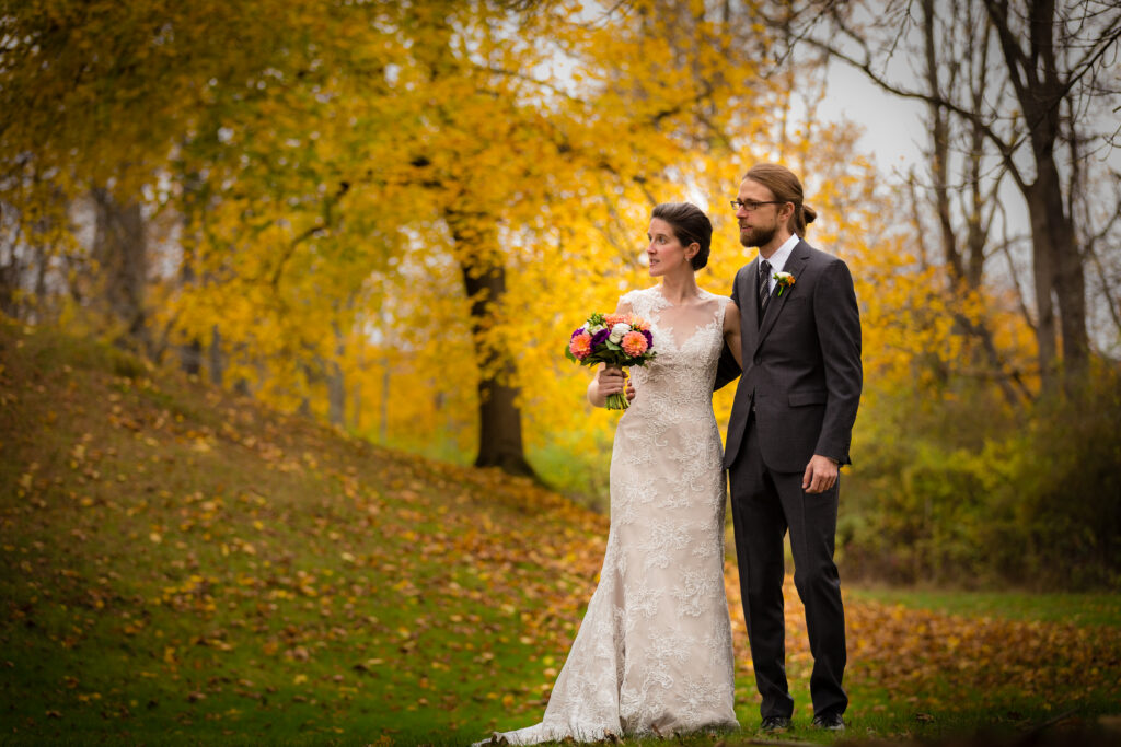 Bride and groom pose among golden autumn trees with a colorful bouquet.