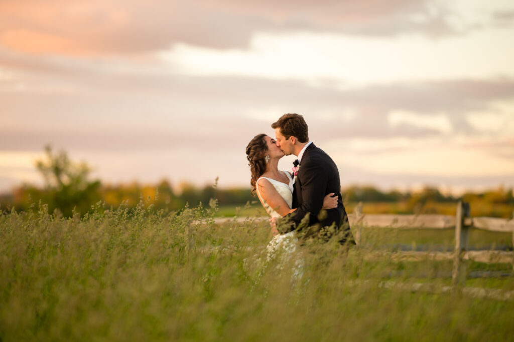 Bride and groom kiss in a grassy field at sunset.