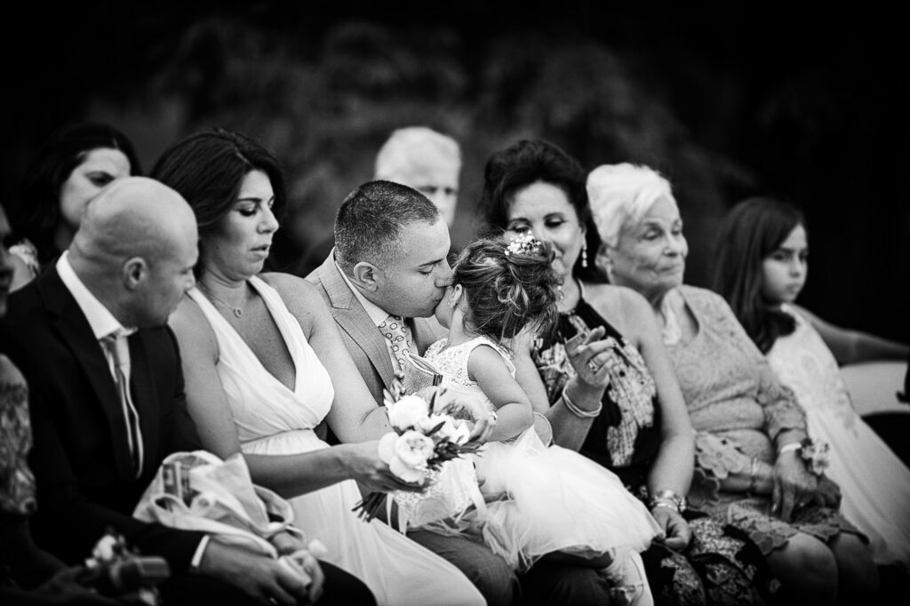 Father kisses a young flower girl seated among wedding guests.