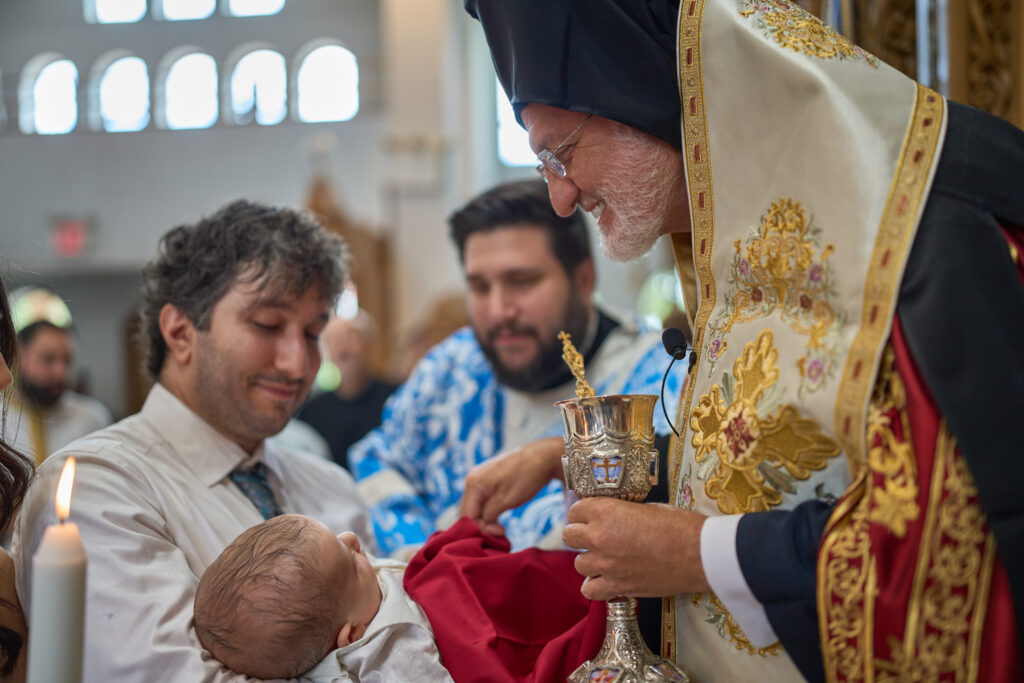 Greek Orthodox priest holds a chalice as a father cradles his infant during baptism.