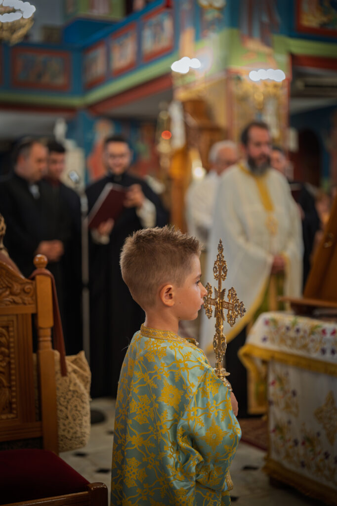 A child holding an ornate cross stands before clergy and chanters.